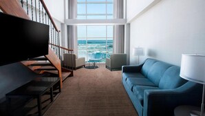A modern living room with a large TV, a blue sofa, a coffee table, and a view of the ocean through large windows. - Niagara Falls Marriott on the Falls (Niagara Falls)