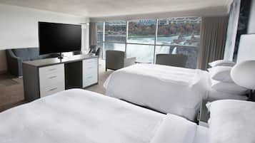 A hotel room with a large bed, a desk with a flat-screen TV, a chair, and a view of a cityscape through large windows.
