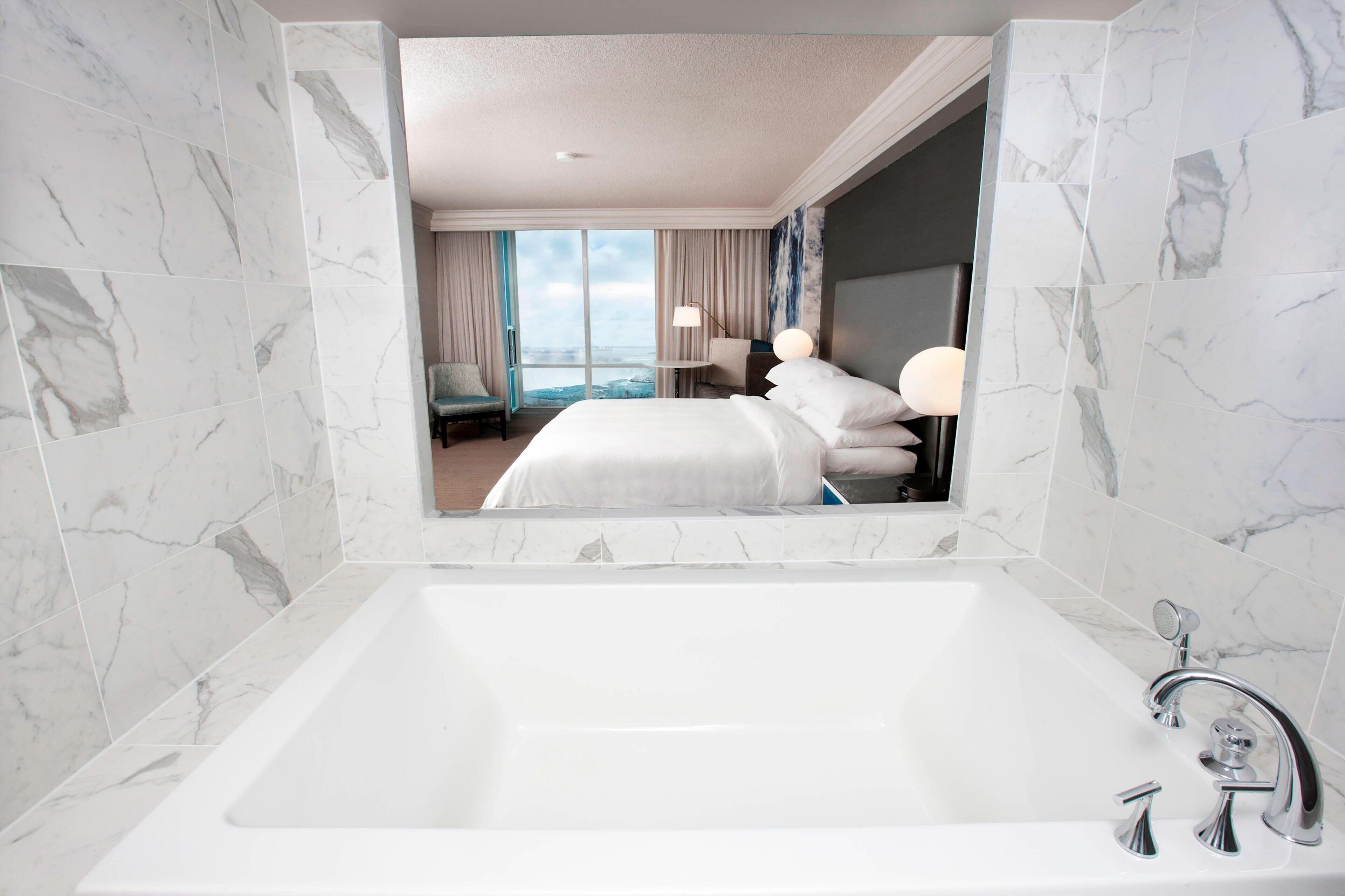 Een moderne hotelbadkamer met een groot wit bad, marmeren wanden en uitzicht op de kamer, inclusief een bed en bureau.
