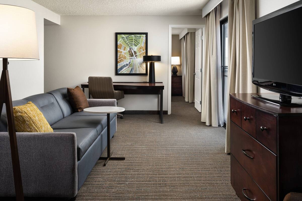 A hotel room with a gray sofa, a small round table, a desk with a lamp, a television, and a wardrobe.