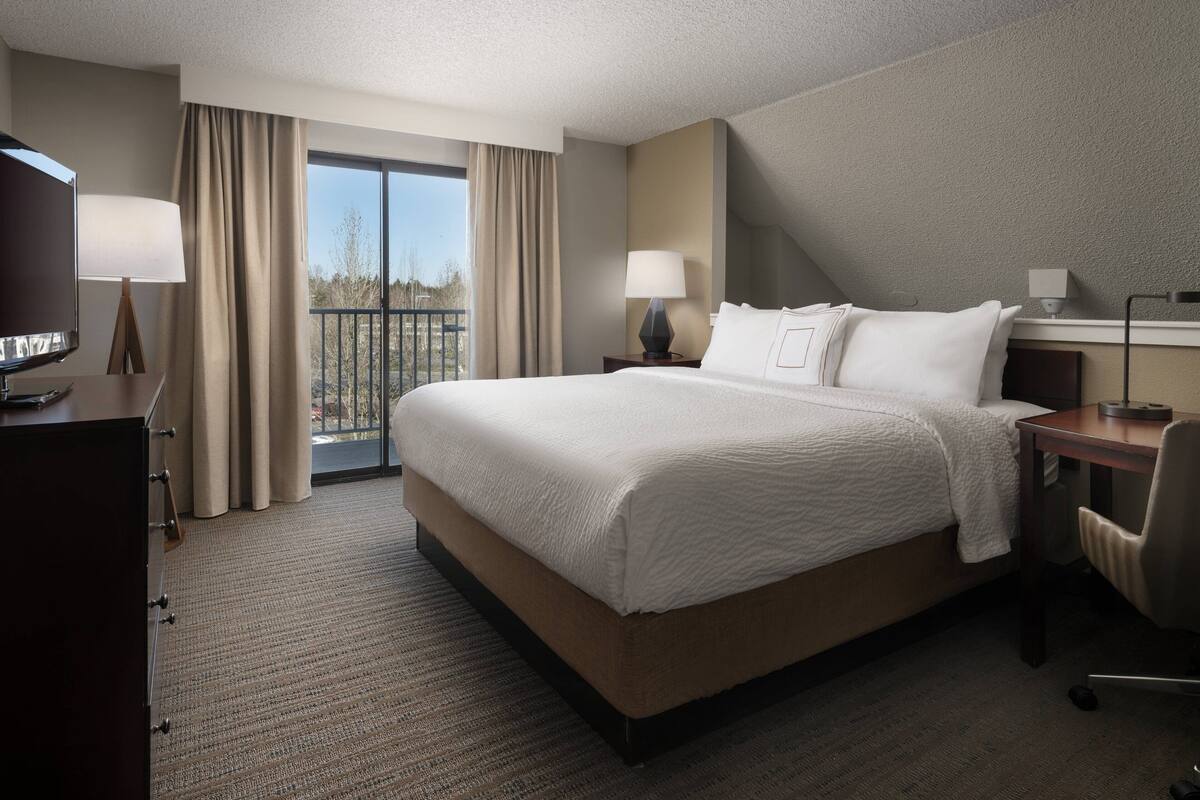 A hotel room with a large bed, two bedside tables, a desk, a chair, a TV, and a balcony with curtains.