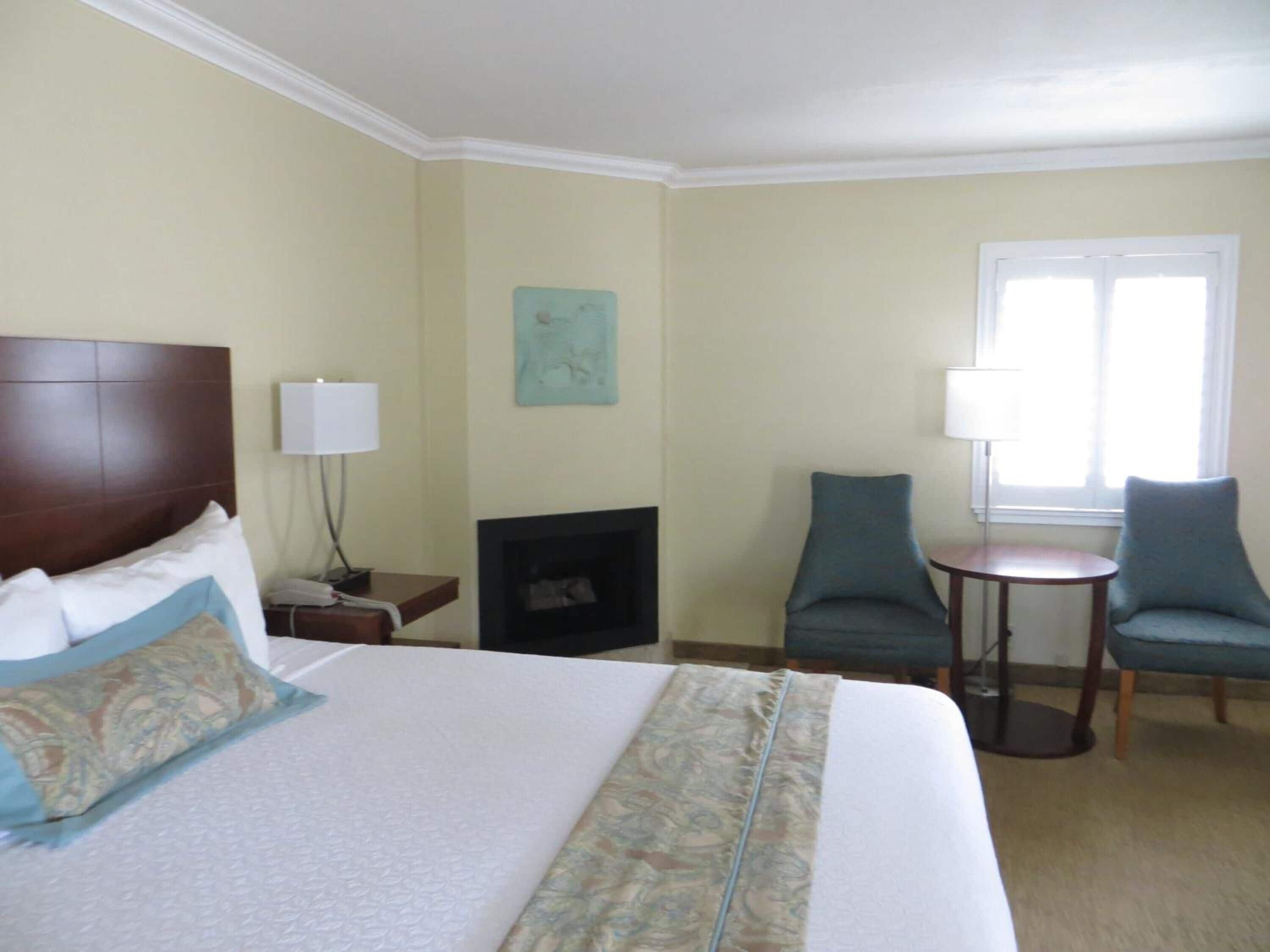 A hotel room with a large bed, two armchairs, a small table, a fireplace, and a window with natural light.
