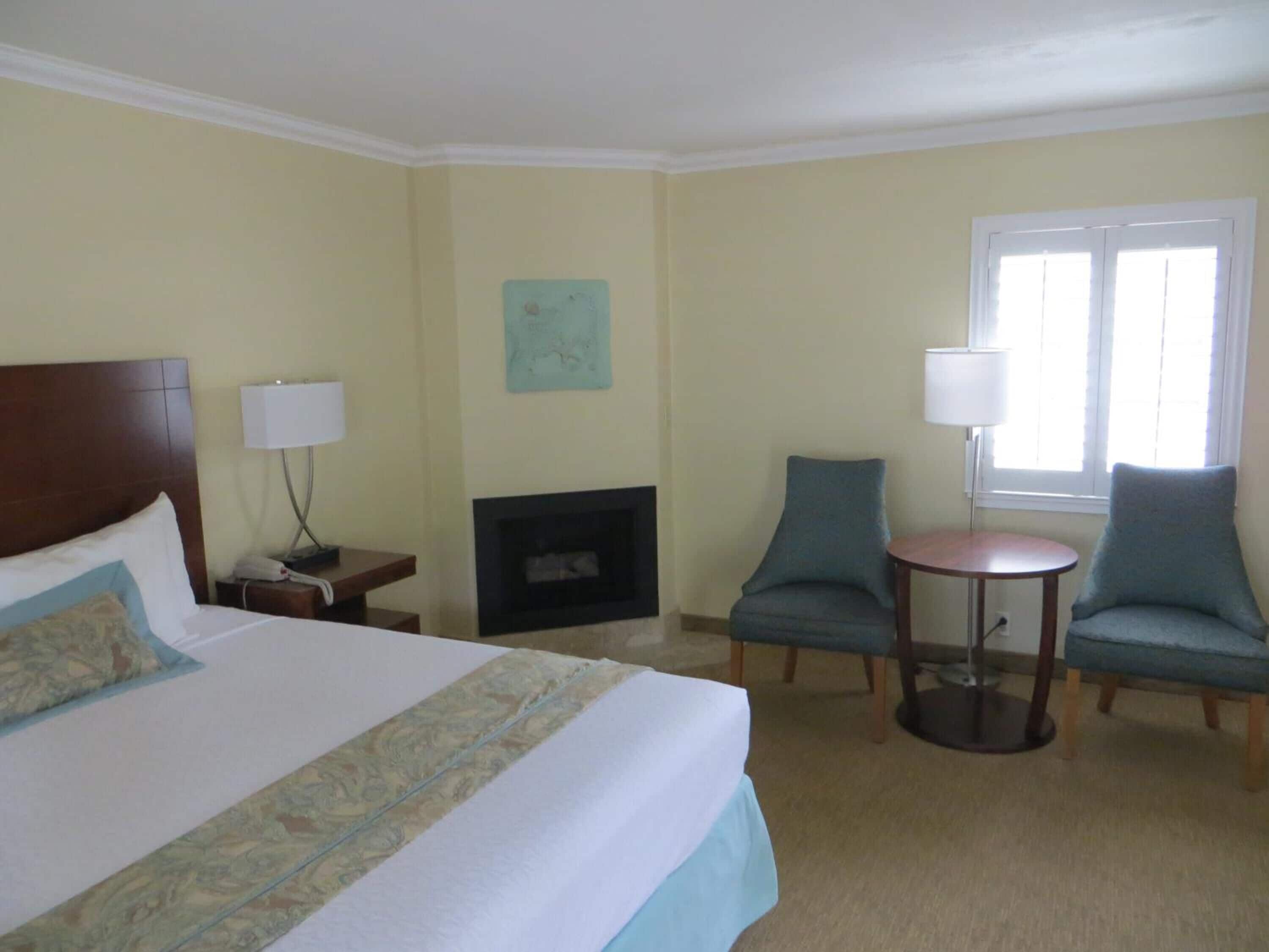Quarto de hotel com uma cama grande, lareira, dois poltronas, uma mesa pequena e uma janela com persianas.