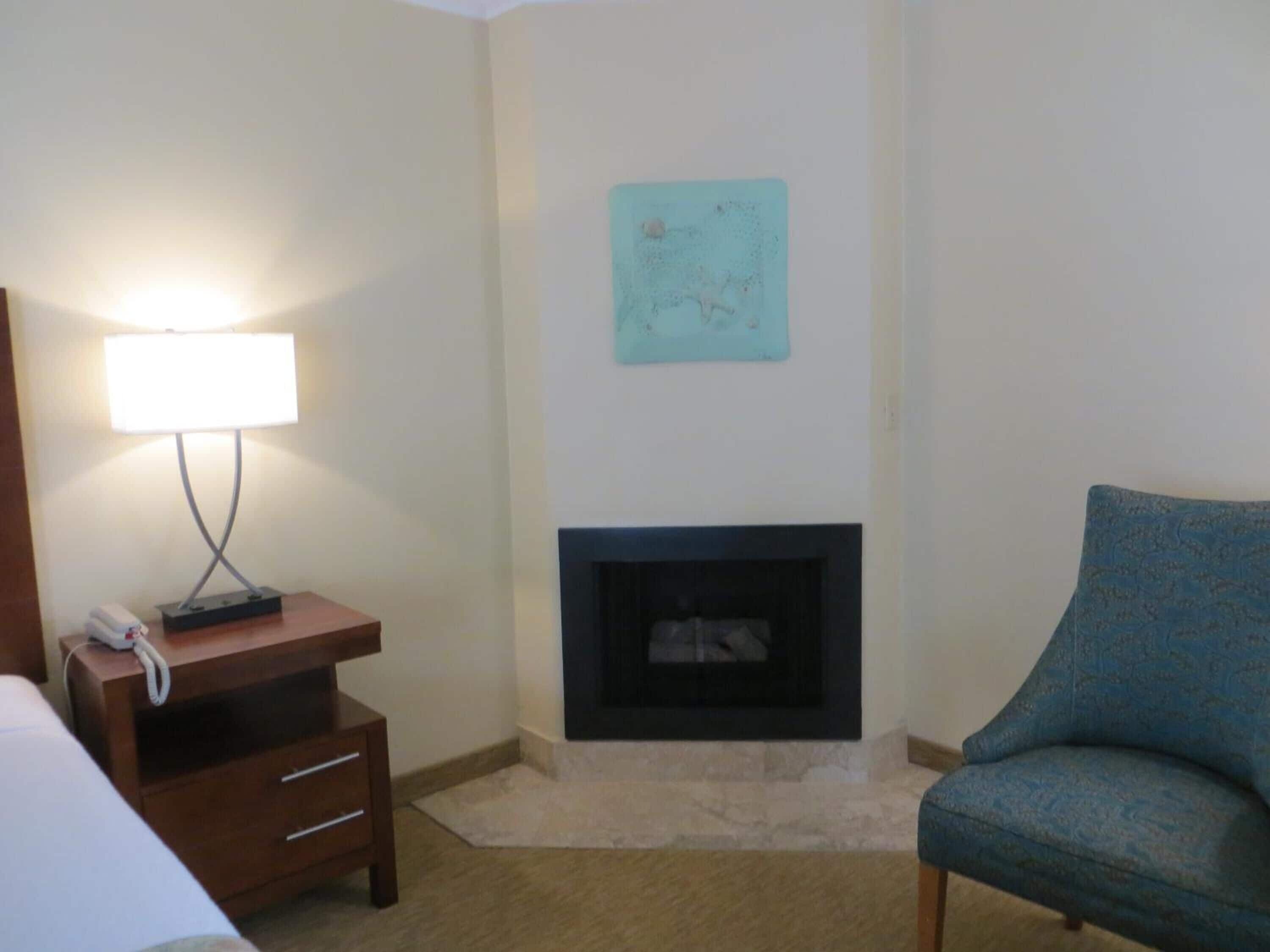A hotel room with a bed, a bedside table with a lamp, a small wooden nightstand, a blue armchair, a fireplace, and a wall map.