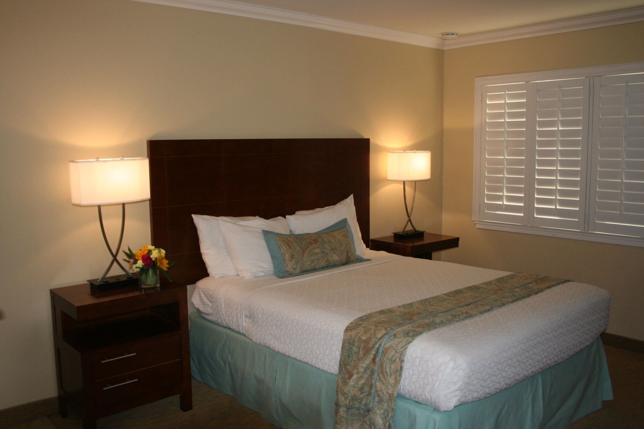 A neatly made bed with a wooden headboard, two bedside tables with lamps, and a window with blinds.