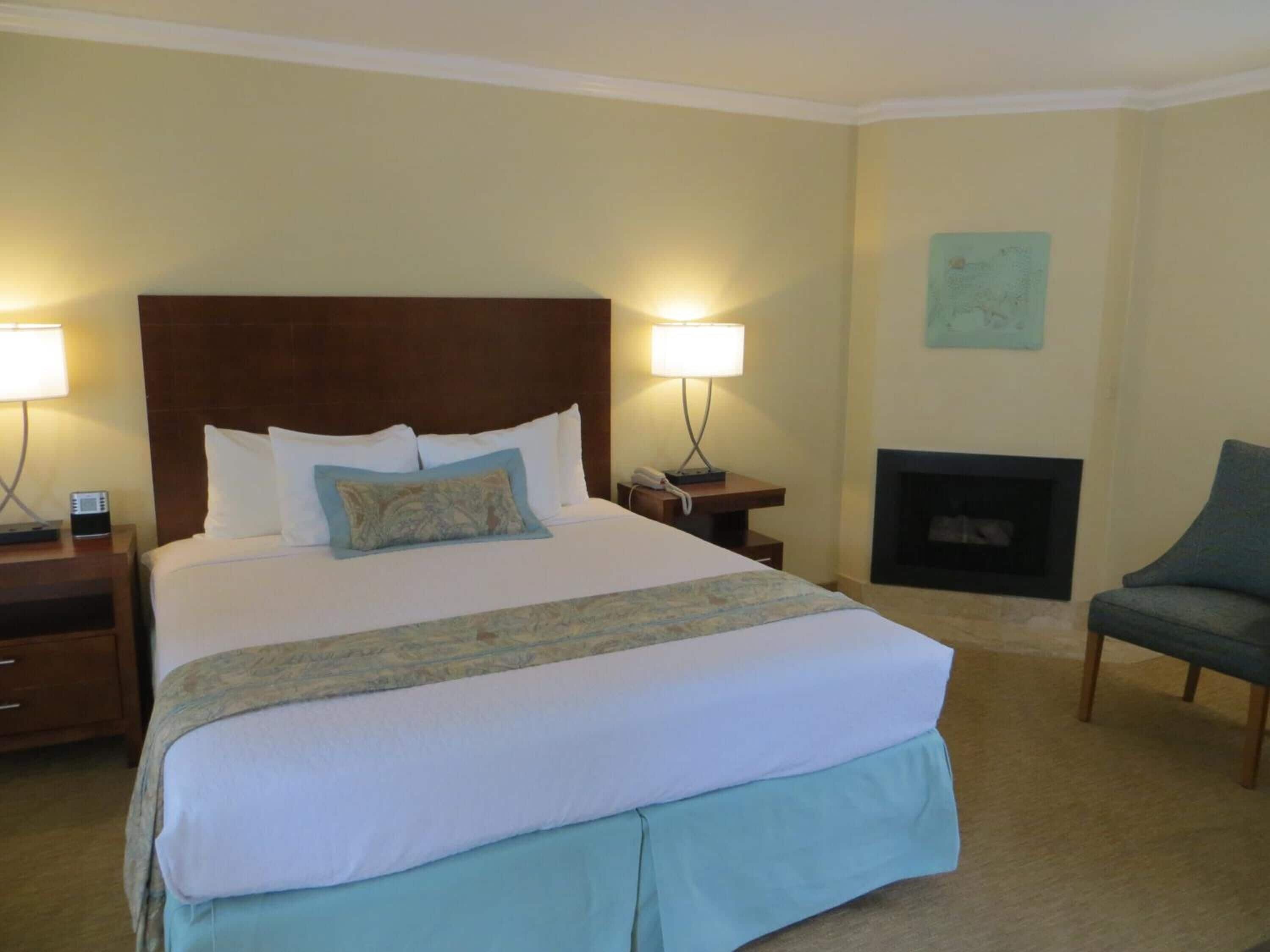 A hotel room with a large bed, two bedside lamps, a nightstand with a phone, a fireplace, and a chair.