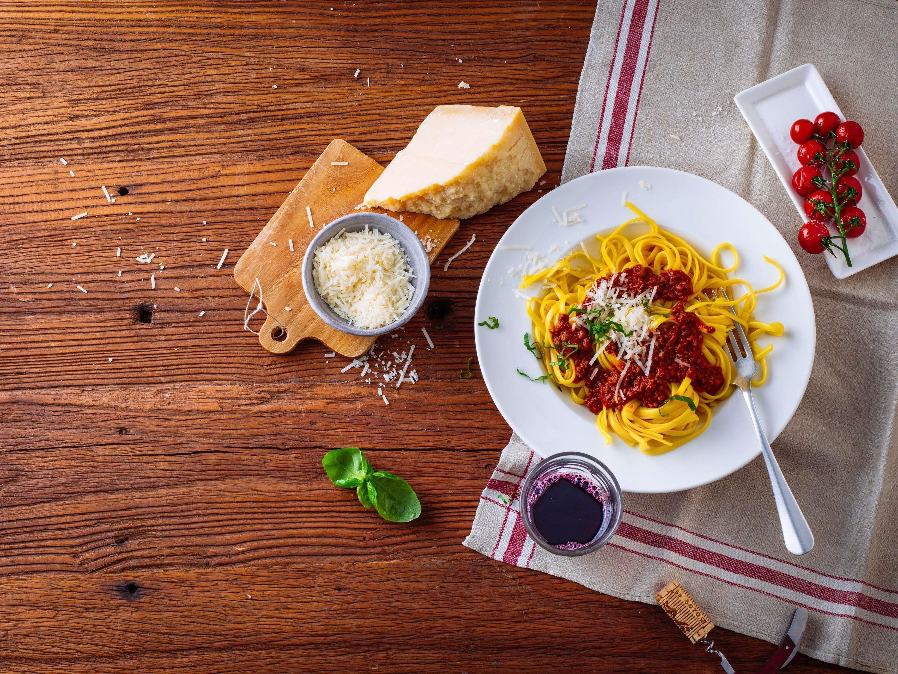 Una mesa de madera con un plato de espagueti, un cuenco de queso rallado, una rebanada de pan, tomates cherry y una copa de vino tinto.
