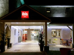 An Ibis Hotels entrance with a glass door, indoor seating area, and a small outdoor patio with chairs and tables.
