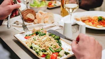 Een eettafel met diverse gerechten, waaronder pasta met groenten, brood en een glas witte wijn.