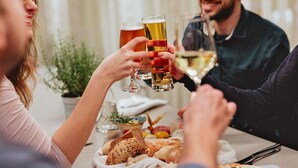 Eine Gruppe von Menschen stoßen mit Bier- und Weingläsern an einem Tisch mit Brot und einer kleinen Schale Obst an.