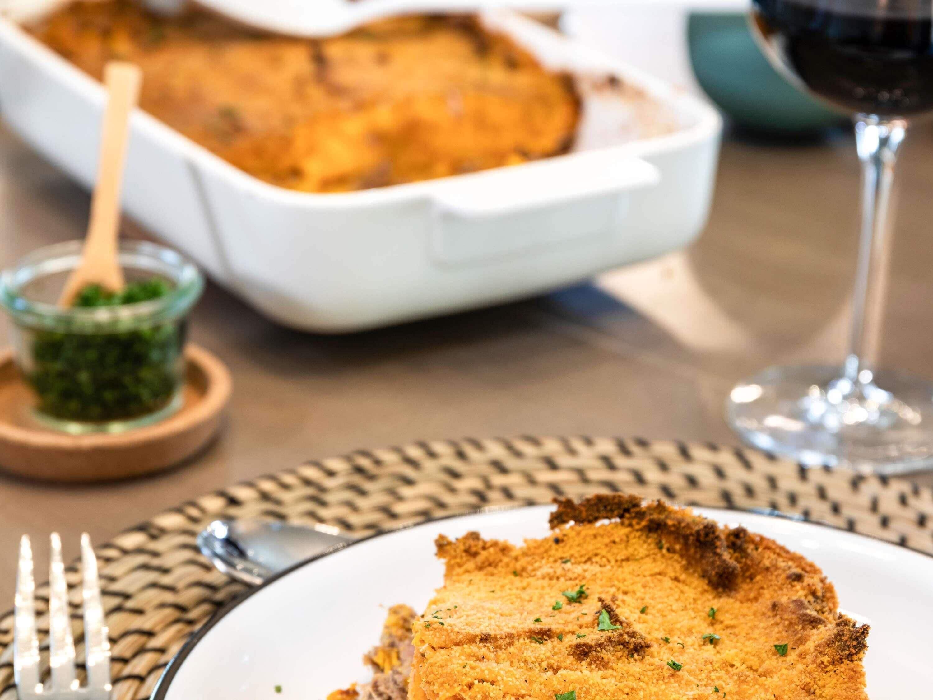 A slice of breaded dish on a plate, a glass of red wine, and a white baking dish in the background.