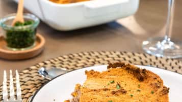 A slice of breaded dish on a plate, a glass of red wine, and a white baking dish in the background.