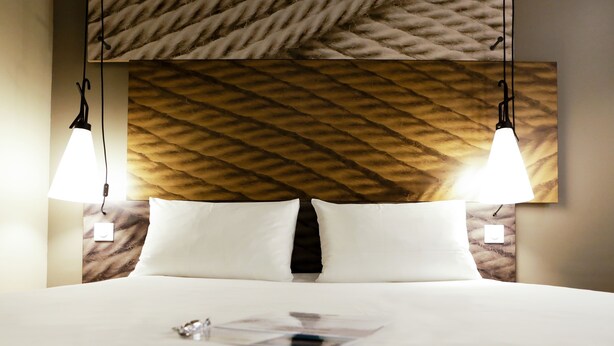 Una habitación de hotel con una cama grande, dos lámparas de noche, un cabecero con textura y una revista sobre la cama.