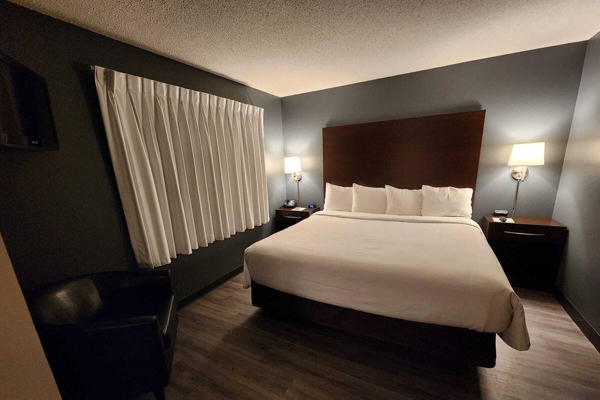 A hotel room with a large bed, two bedside tables with lamps, a chair, and a trash bin.