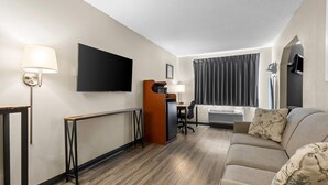 Habitación de hotel con sofá, televisión, escritorio con silla, lámpara y ventana con cortinas.