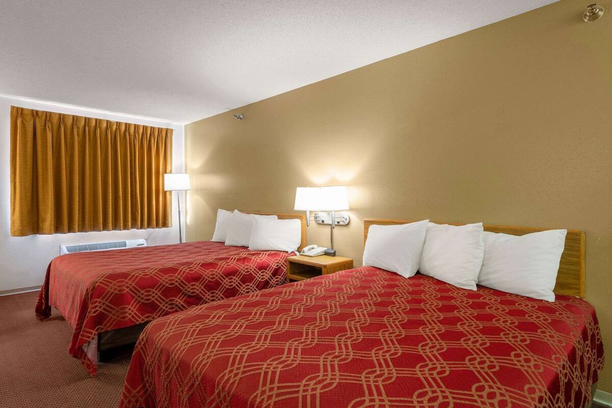 A hotel room with two beds, a window with curtains, a bedside table with a lamp, and a wall-mounted phone.