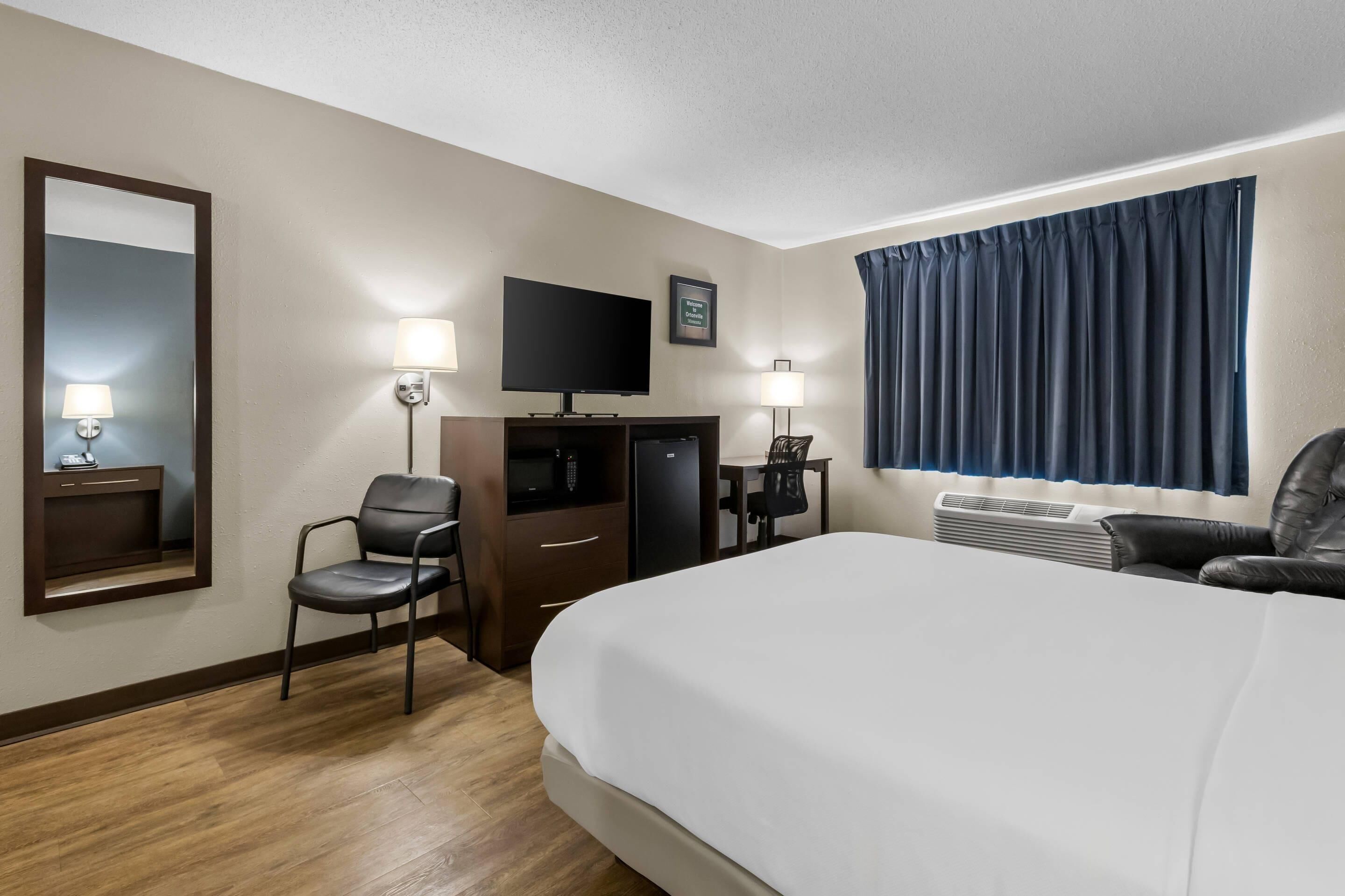 Habitación de hotel con una cama grande, un escritorio con silla, un televisor, un espejo y una ventana con cortinas.