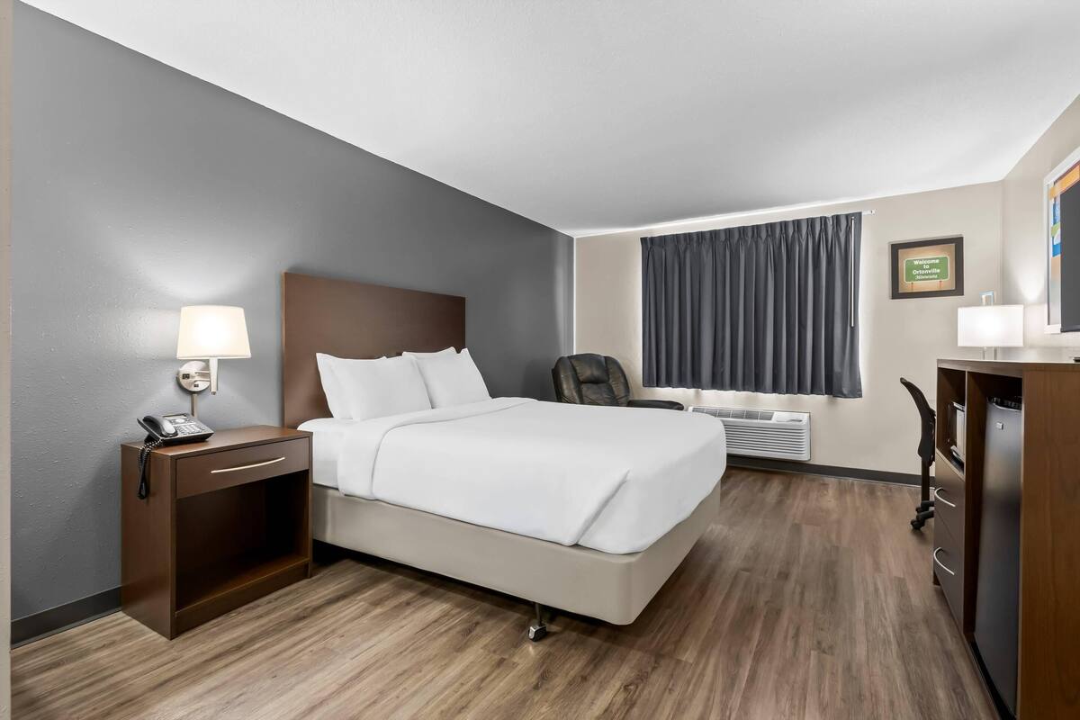 A hotel room with a bed, bedside table with a lamp, a desk with a chair, a window with curtains, and a wall-mounted TV.