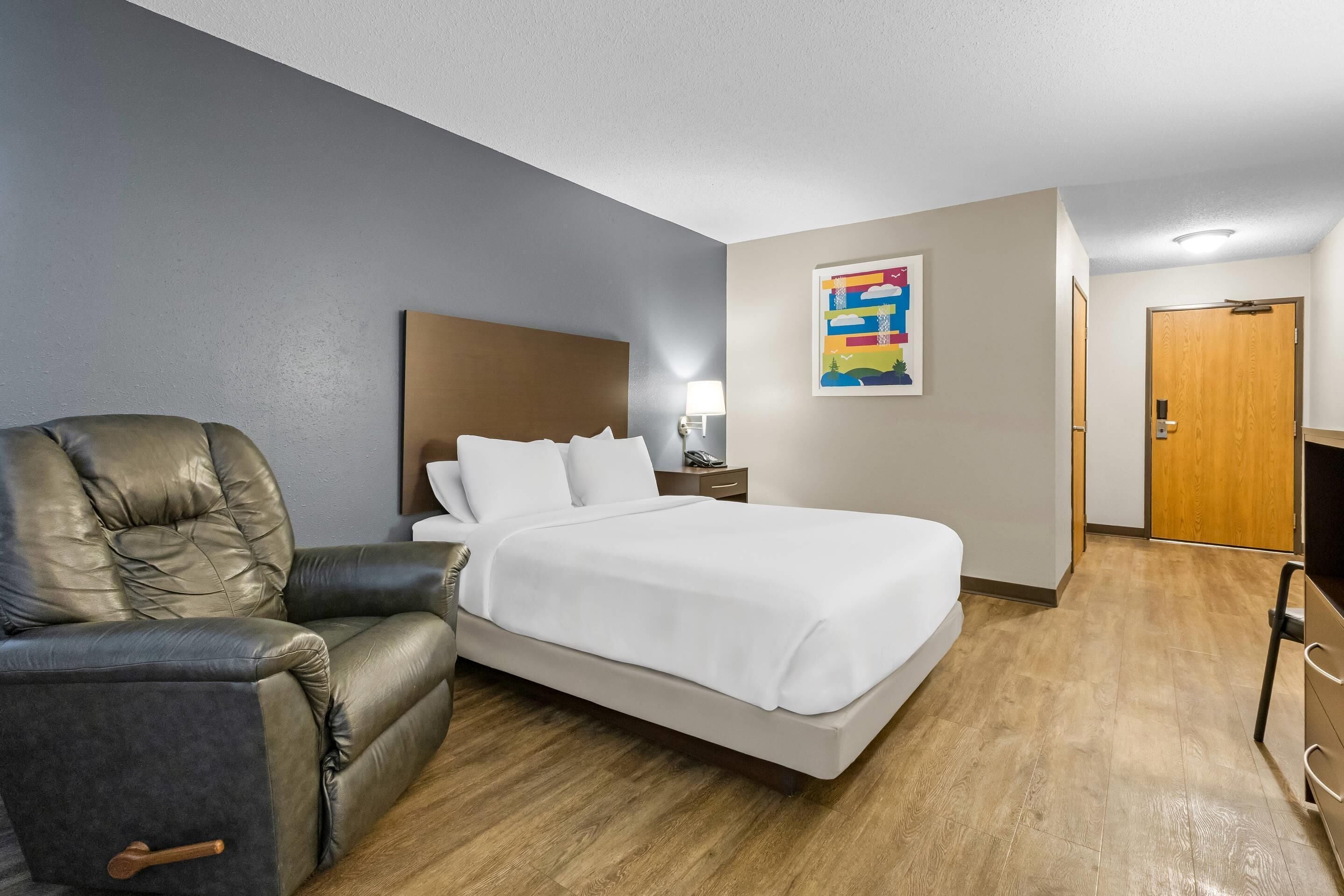 A hotel room with a bed, a leather armchair, a nightstand, a lamp, a wooden door, and a colorful artwork on the wall.