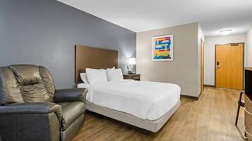 A hotel room with a bed, a leather armchair, a nightstand, a lamp, a wooden door, and a colorful artwork on the wall.