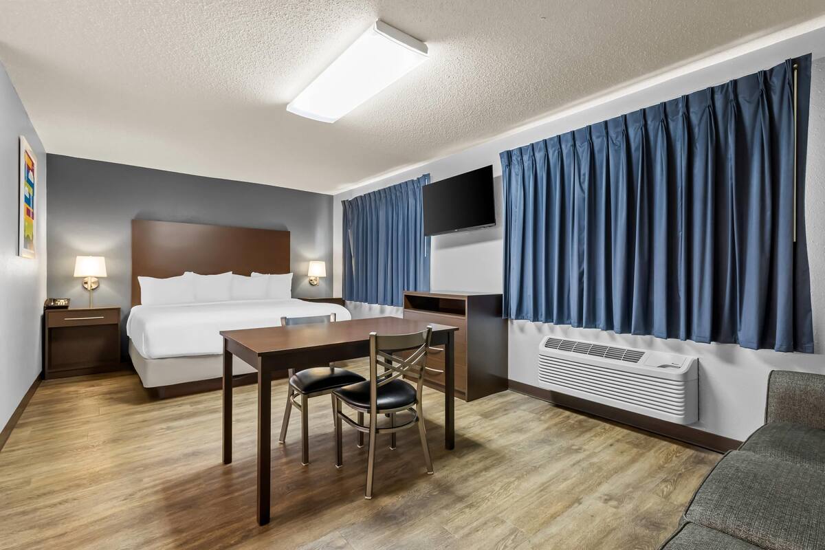 A hotel room with a bed, desk, chair, TV, and air conditioning unit.