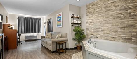 A modern hotel room with a wooden floor, a large bathtub, a sofa, a small table, a potted plant, and a wall-mounted shelf with towels.