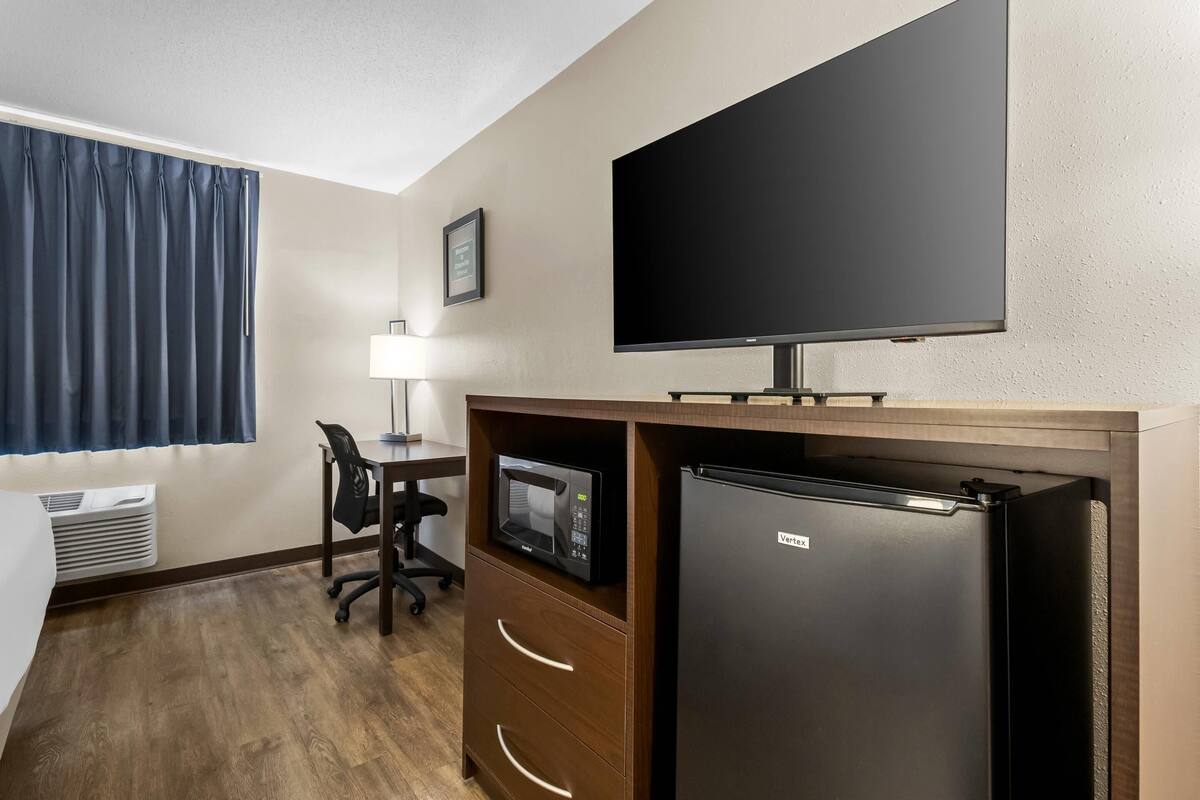 A hotel room with a flat-screen TV, a refrigerator, a microwave, a desk with a chair, and a window with curtains.