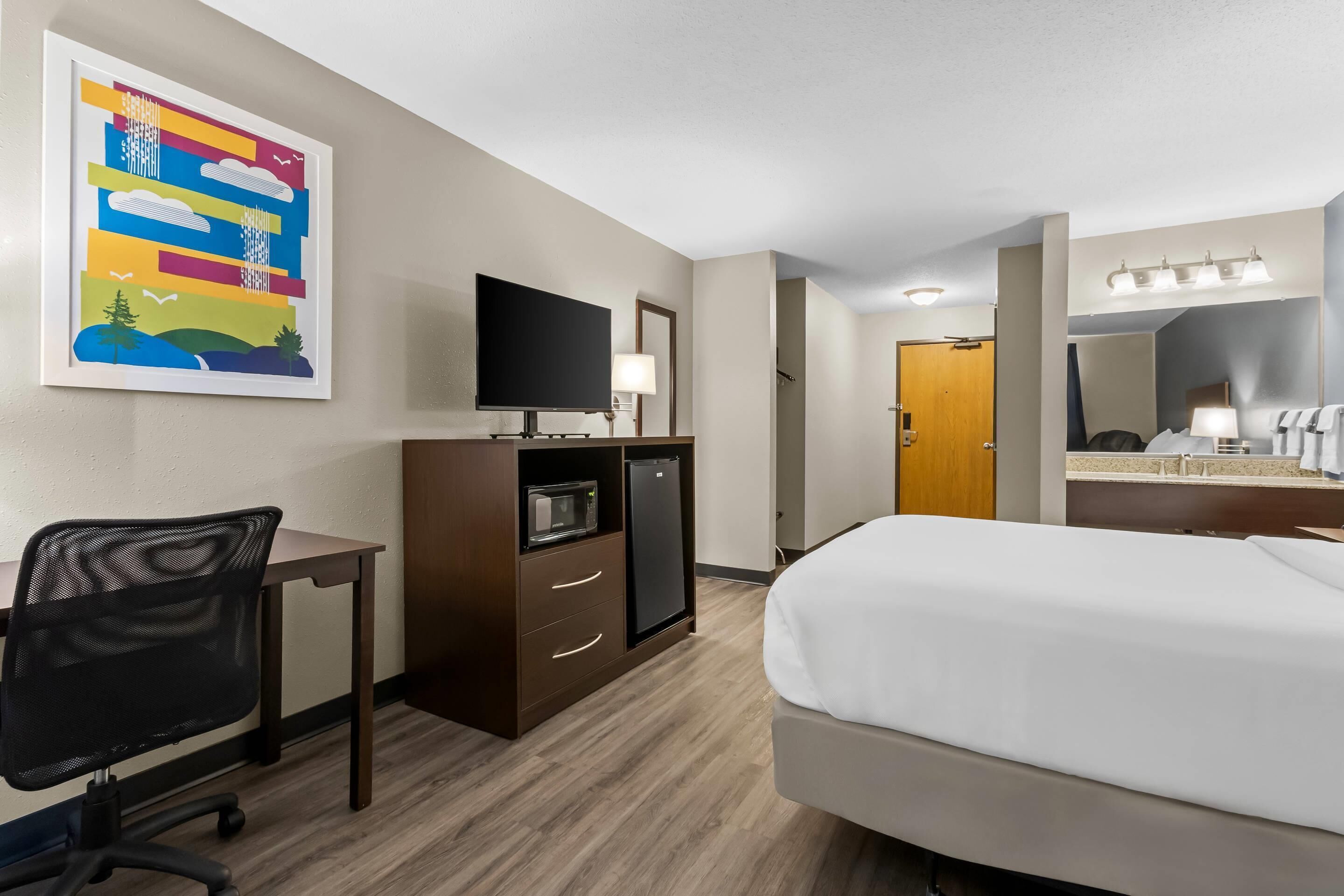 A hotel room with a bed, desk, chair, TV, microwave, refrigerator, and a painting on the wall.