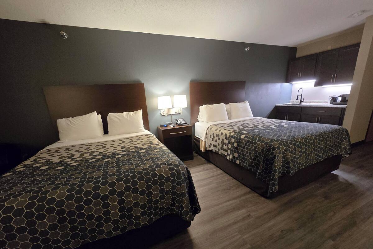 A hotel room with two beds, a small desk with a lamp, a kitchenette, and a bathroom.