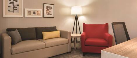 Una habitación de hotel con un sofá color beige, un sillón rojo, una mesita redonda pequeña y una lámpara de pie.