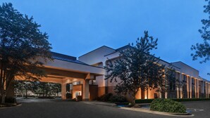 A modern hotel with a covered entrance, a parking lot, and surrounding greenery.