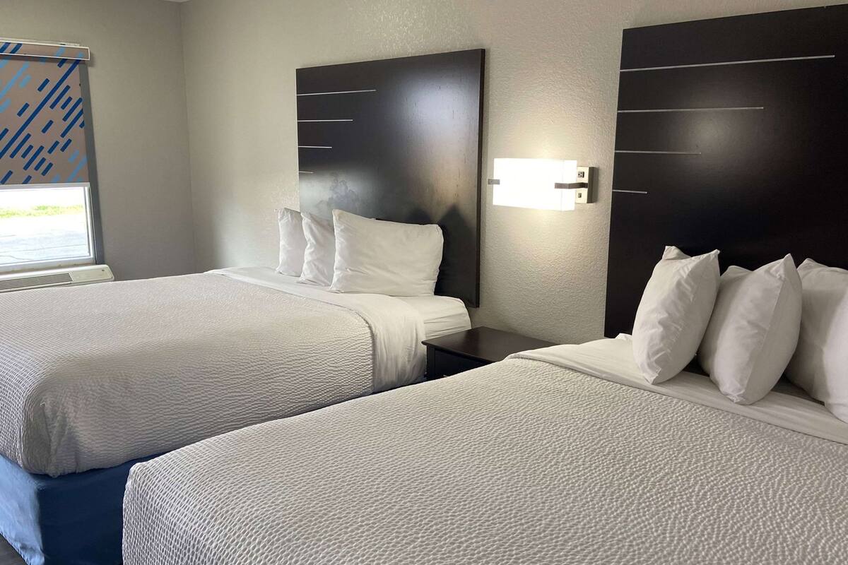 A hotel room with two beds, a large window with blinds, and a wall-mounted light fixture.
