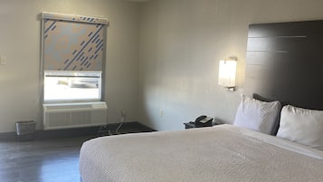 Habitación de hotel con una cama grande, una mesita de noche con teléfono, una ventana con persianas, un equipo de aire acondicionado y una lámpara de pared.