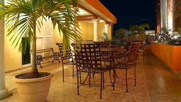 An outdoor patio with a dining area, including a large table and chairs, surrounded by potted plants and a palm tree.