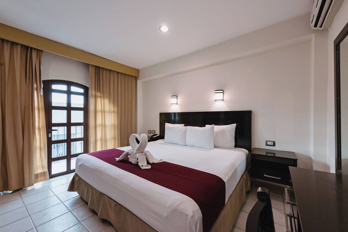 A hotel room with a large bed, a desk, a chair, a window with curtains, and a wall-mounted air conditioning unit.