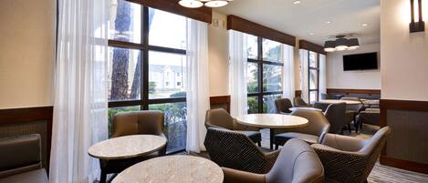 Un comedor de hotel con mesas redondas, asientos acolchados, ventanas amplias con cortinas y un televisor de pantalla plana en la pared.