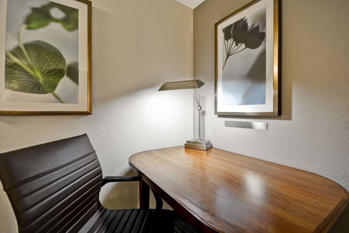 Una habitación de hotel con un escritorio de madera, una silla, una lámpara y dos impresiones enmarcadas de motivos botánicos en la pared.
