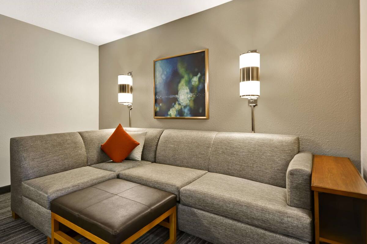 Una habitación de hotel moderna con un sofá seccional gris, una mesa de centro de madera y dos lámparas fijadas a la pared.