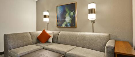 A modern hotel room with a grey sectional sofa, a wooden coffee table, and two wall-mounted lamps.