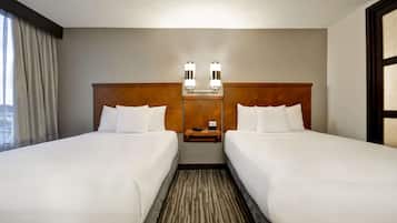 Quarto de hotel com duas camas, cabeceira de madeira, mesa de cabeceira e uma janela com cortinas.