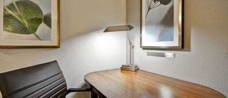 A hotel room with a wooden desk, a chair, a lamp, and two framed botanical prints on the wall.