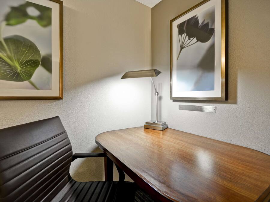 Une chambre d’hôtel avec un bureau en bois, une chaise, une lampe et deux reproductions botaniques encadrées accrochées au mur.