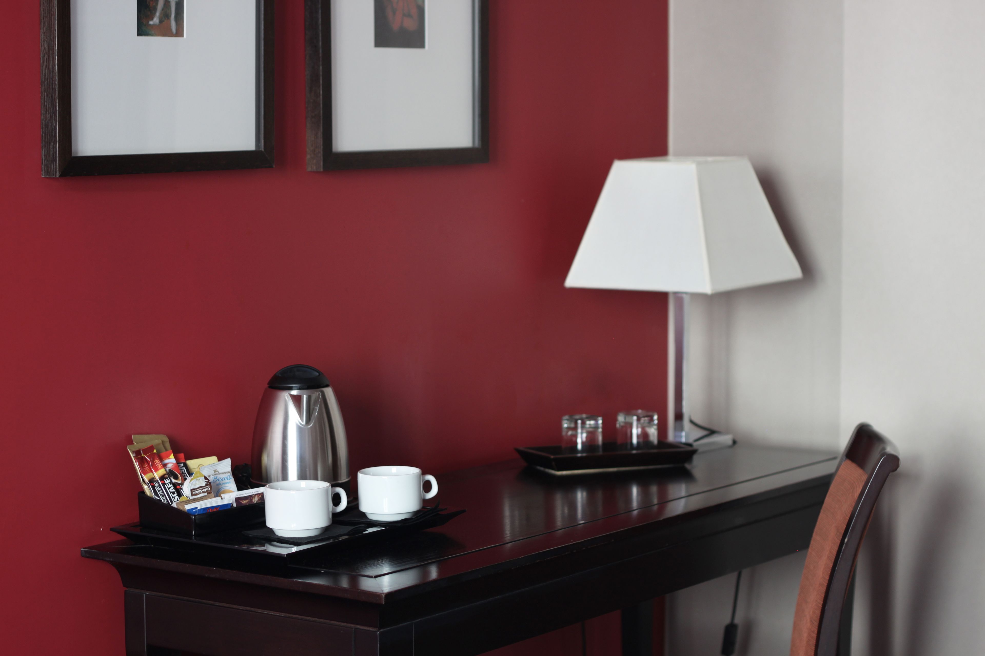 A hotel room with a dark wooden desk, a white coffee cup, a white mug, a silver coffee pot, a tray with glasses, and a lamp.