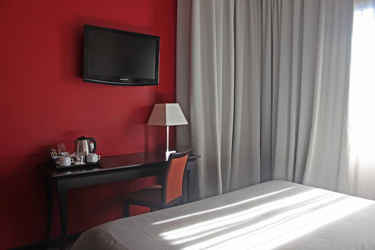 A hotel room with a bed, a desk with a lamp, a television, and a window with curtains.