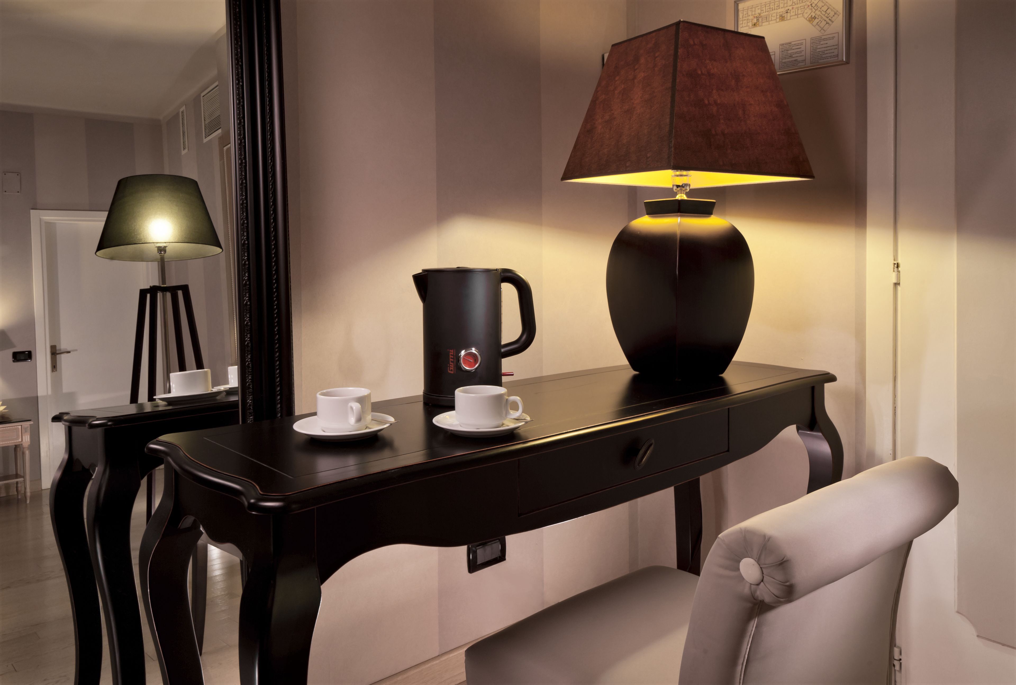 A modern hotel room with a dark wooden desk, a white chair, a lamp, a coffee maker, and a cup of coffee.