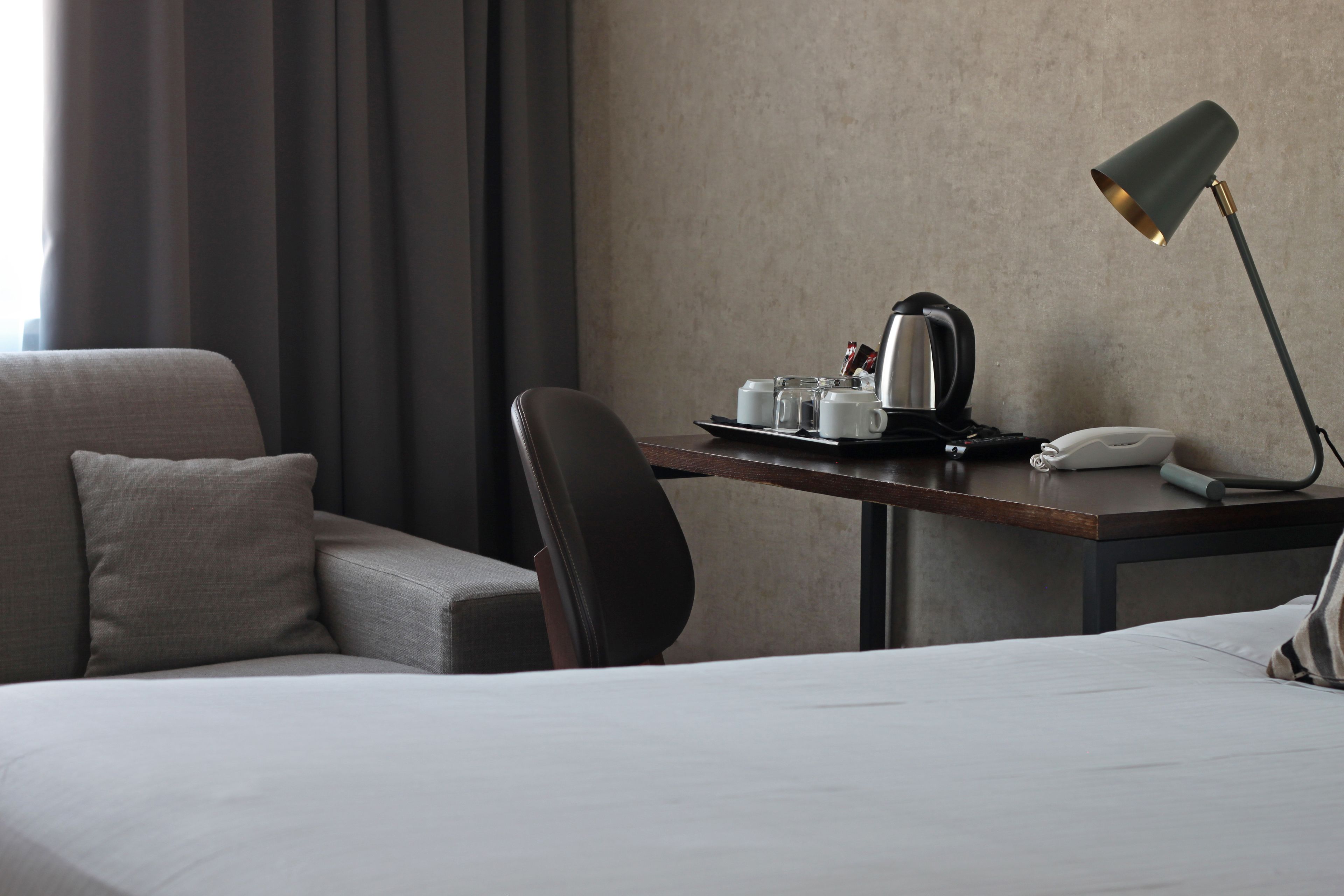 Quarto de hotel com cama, escrivaninha com luminária, cadeira, cafeteira e bandeja com chávenas e bule.