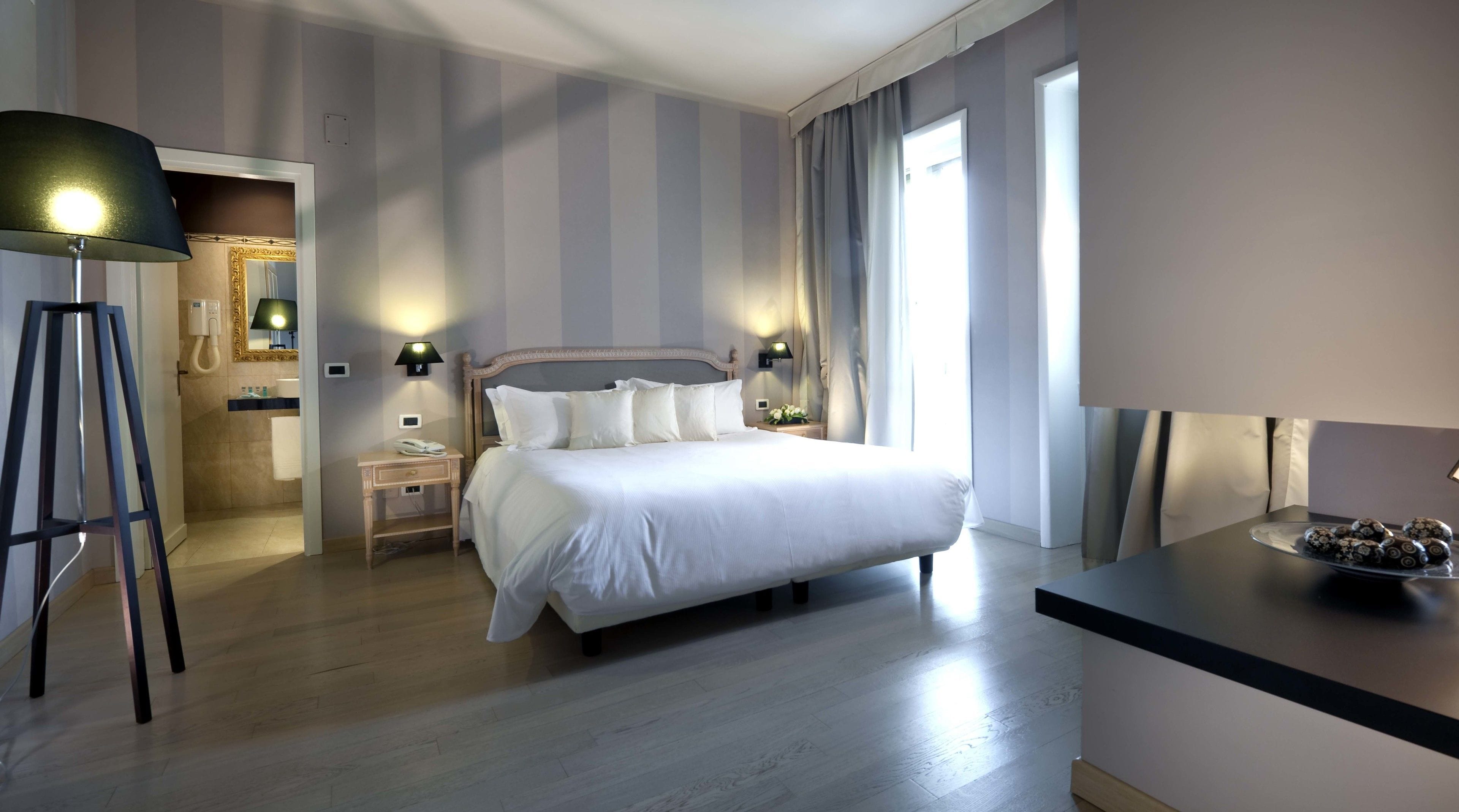 A hotel room with a large bed, bedside lamps, a desk with a lamp, a mirror, and a view of the outdoors.