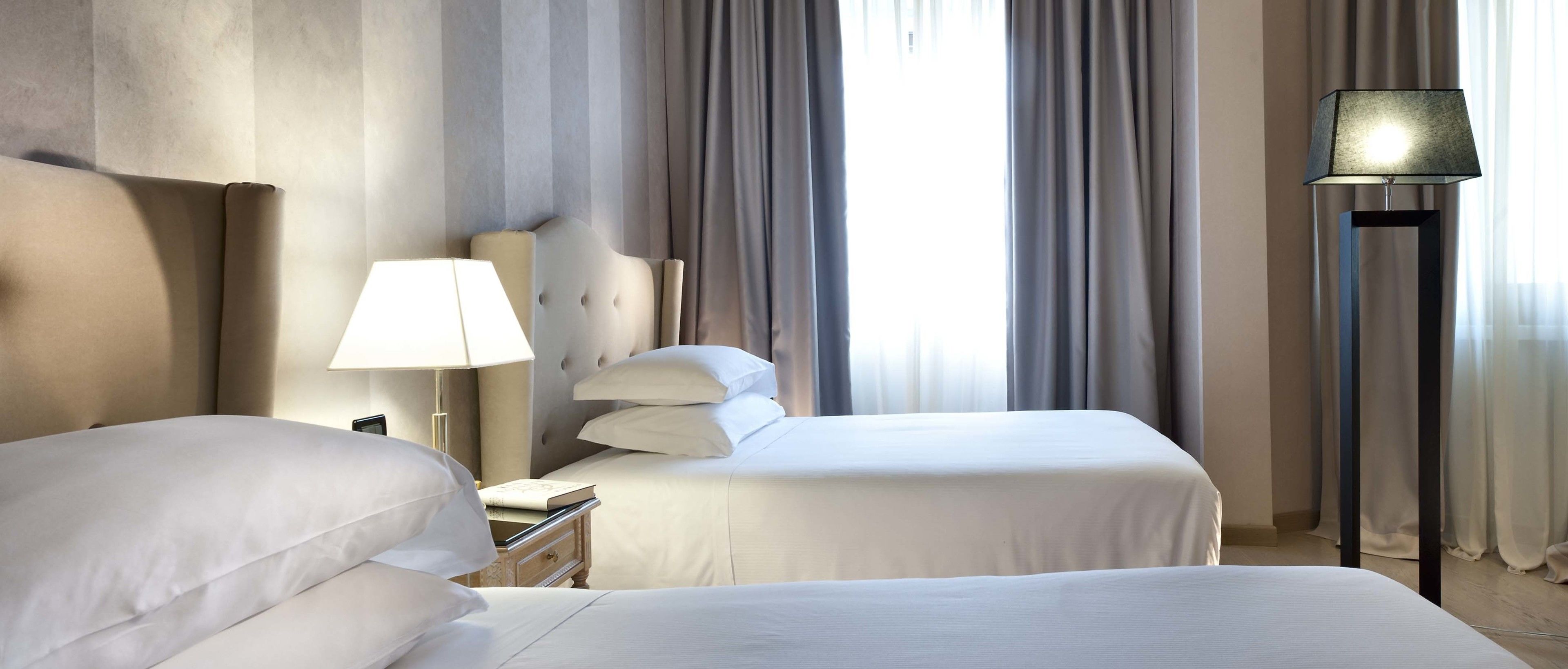 A hotel room with two beds, a bedside table with a lamp, and a view of the outdoors through large windows.