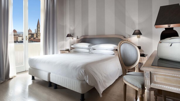 A hotel room with a large bed, a chair, a desk, and a view of a cityscape through a balcony. - c-hotels Ambasciatori (Florence)