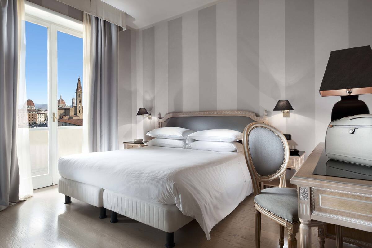 A hotel room with a large bed, a chair, a desk, and a view of a cityscape through a balcony.